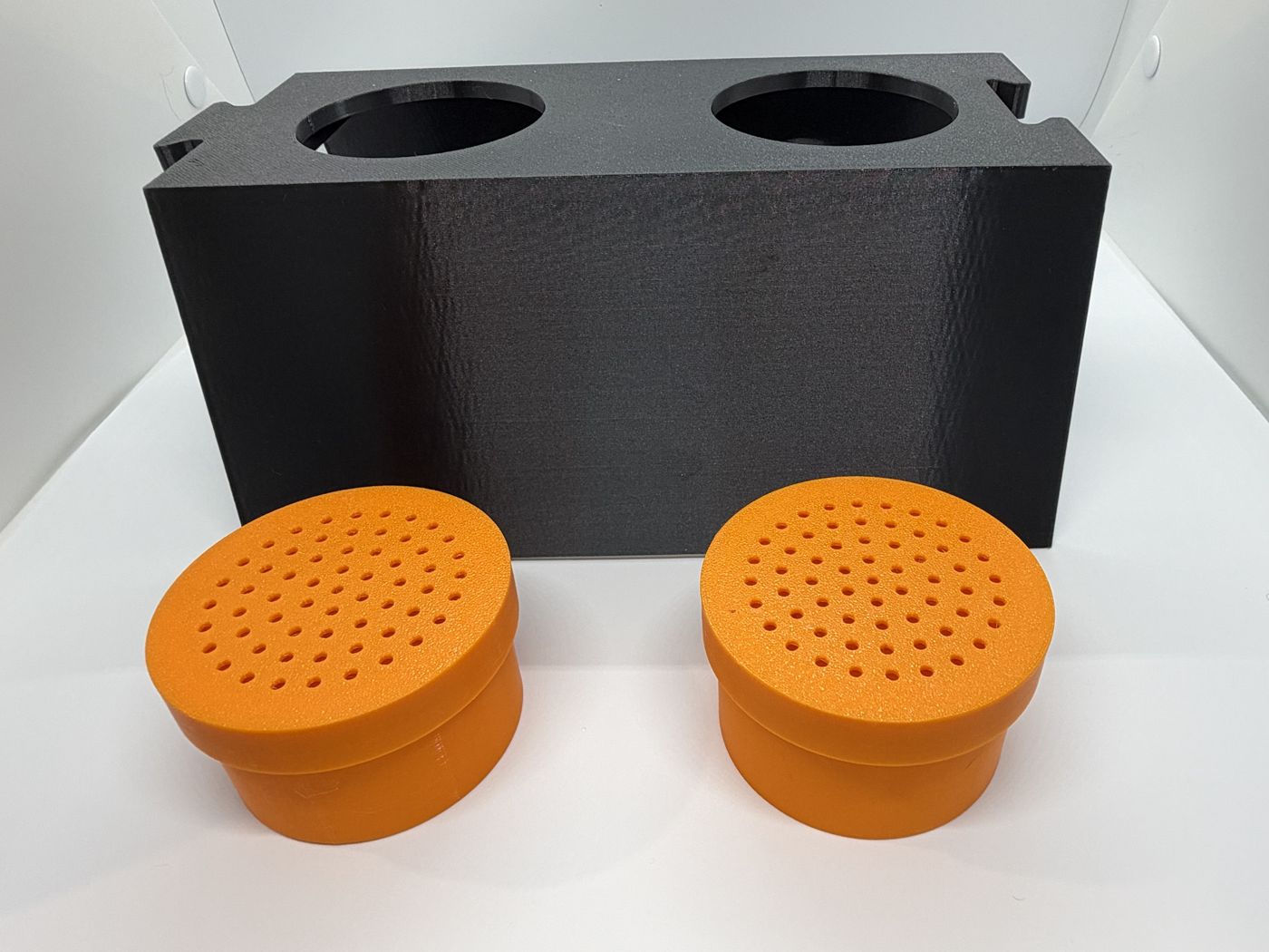 A black two-hole box with two orange perforated scent pods in front of it